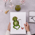 Person holding a card with a cartoon kappa illustration on a table with a glass of green liquid and flowers.
