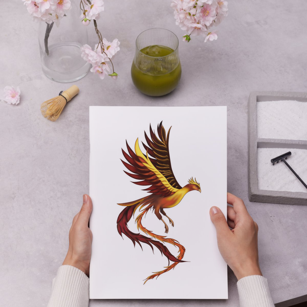 Person holding a card with a phoenix design on a table with a glass of green liquid and flowers.